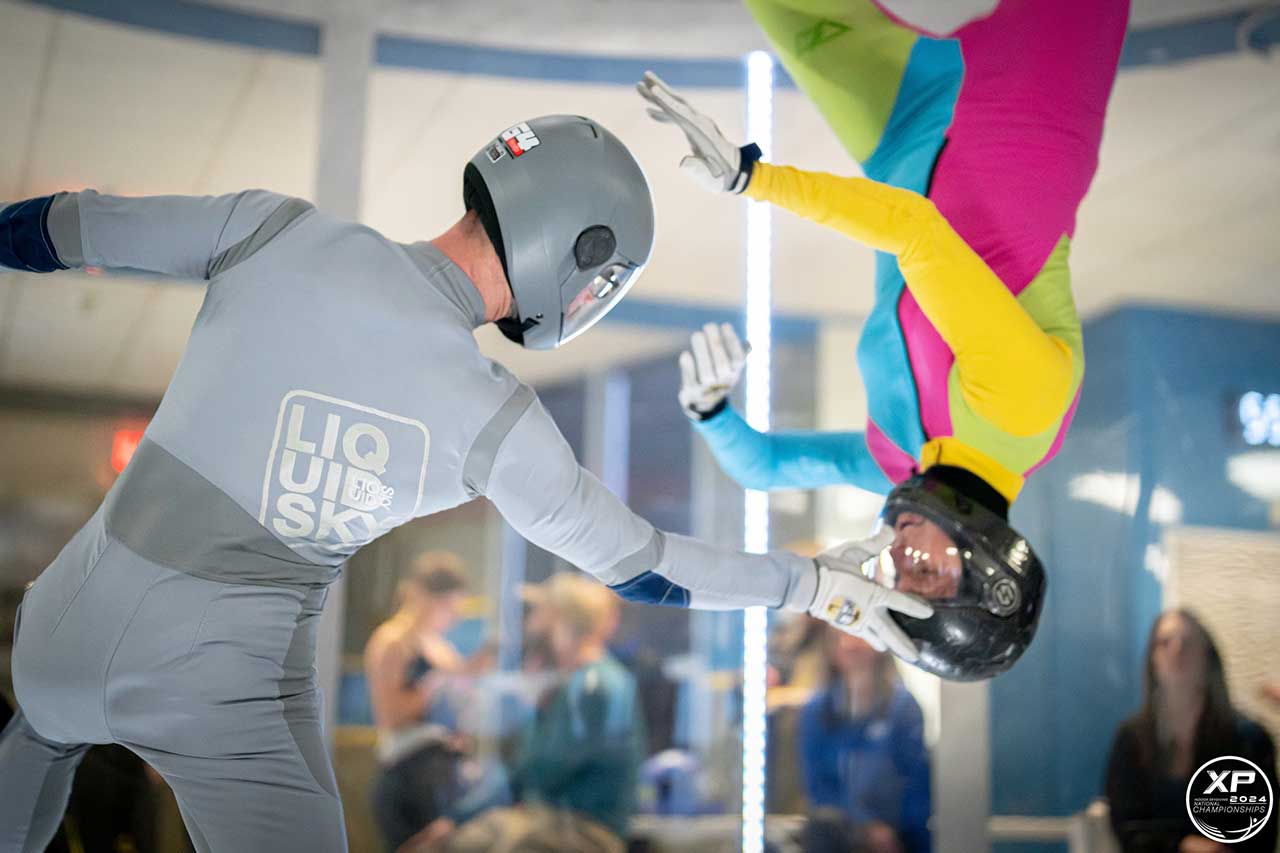 Indoor skydiver in a gray suit and helmet touching the head of another upside down skydiver