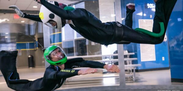 Two indoor skydivers in a white and green helmet fly and smile together.
