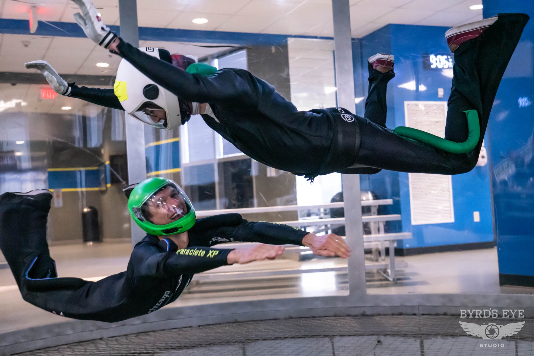 Two indoor skydivers fly in the tunnel with full face green and white helmets.