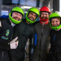 Group of smiling indoor skydivers wearing green and red helmets posing for the camera.