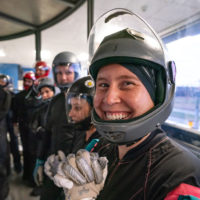 Excited indoor skydiver smiles at the camera with her helmet's visor up and hands clasped.