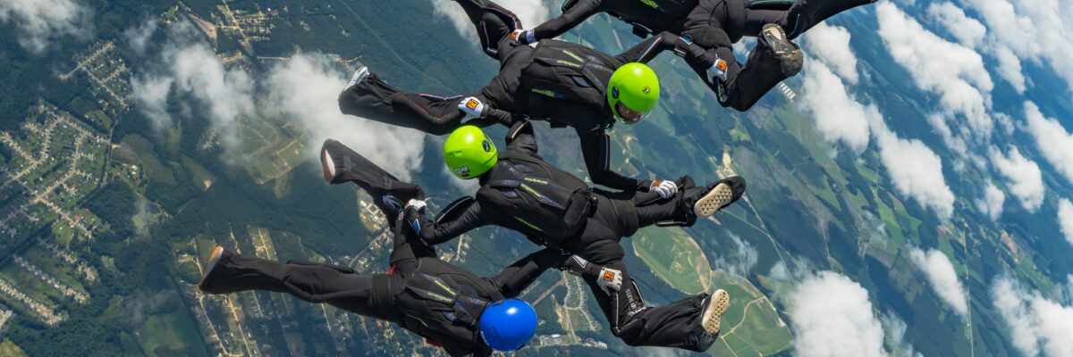 Four skydivers in freefall in black jumpsuits and green and blue helmets.