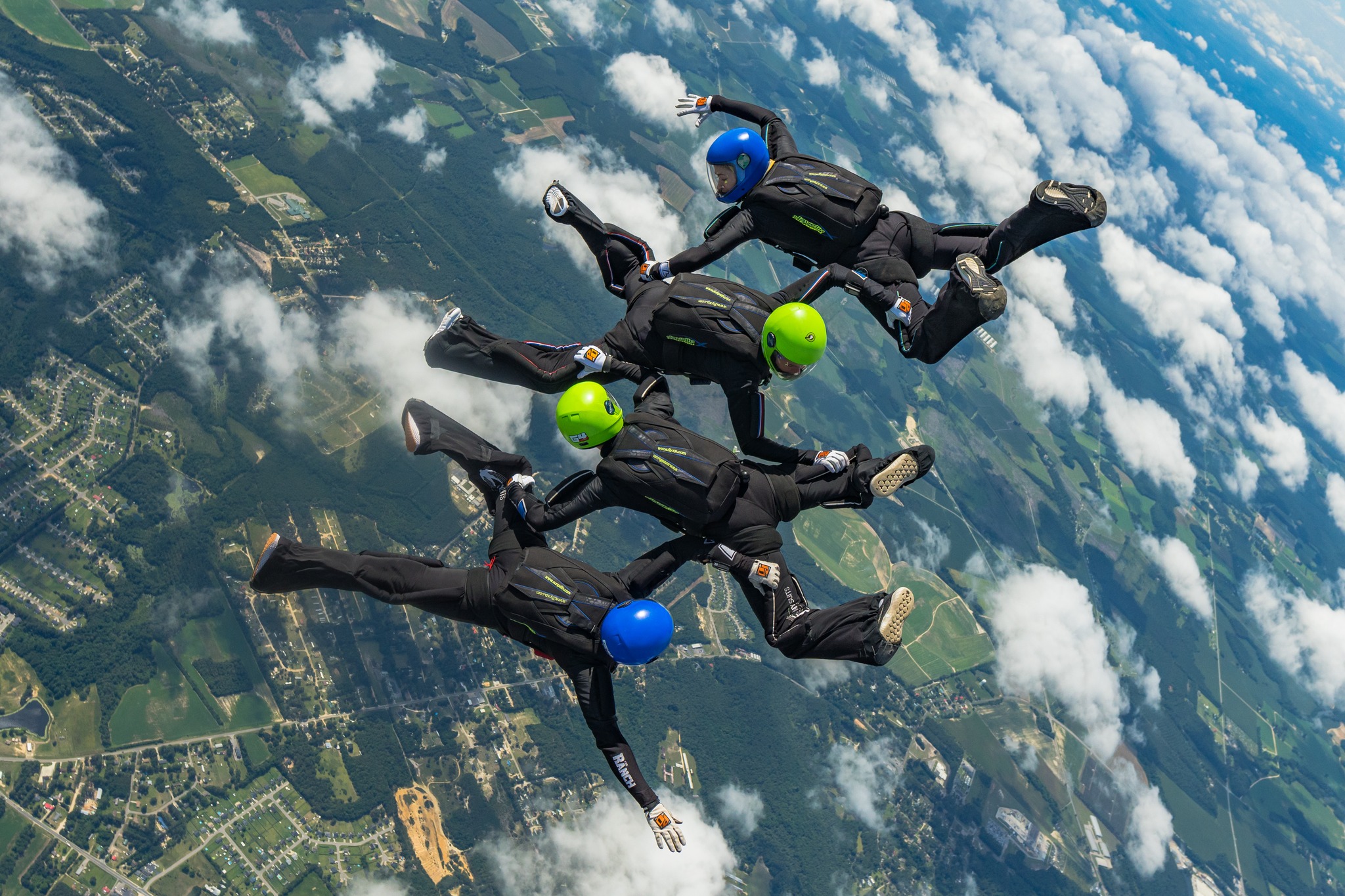 Four skydivers in freefall in black jumpsuits and green and blue helmets.