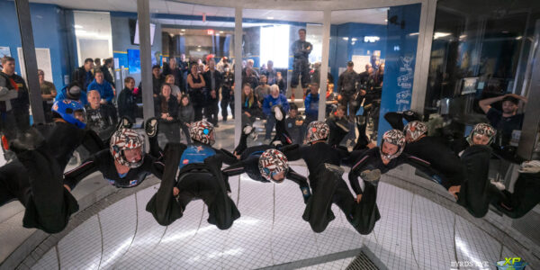 Eight fliers in the wind tunnel wearing black jumpsuits and patriotic helmets side by side.